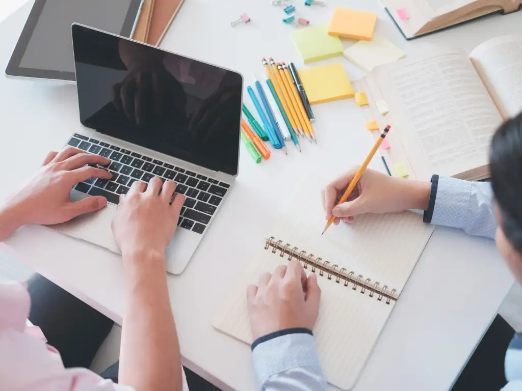 Dua orang menulis di buku catatan dan mengetik di laptop
